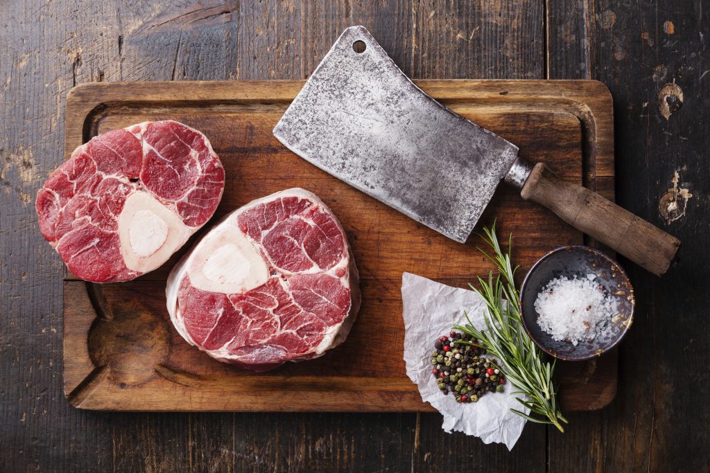 Fresh Osso Buco surrounded by a large butcher knife, peppercorns, rosemary and sea salt on a brown cutting board