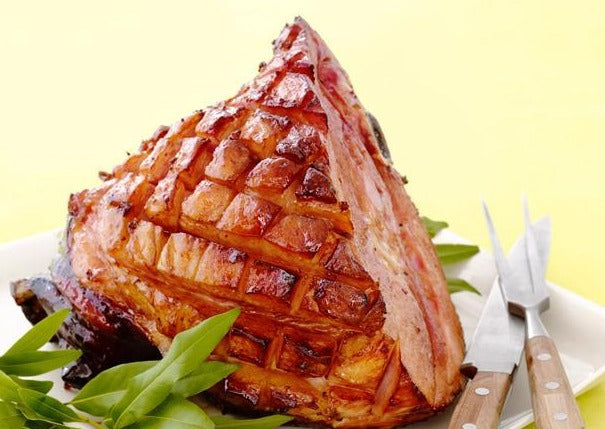 Glazed, bone-in ham with butcher knife and fork against a light yellow background
