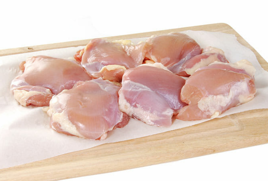 Six fresh bone-in chicken thighs sitting on white parchment paper on a wooden cutting board