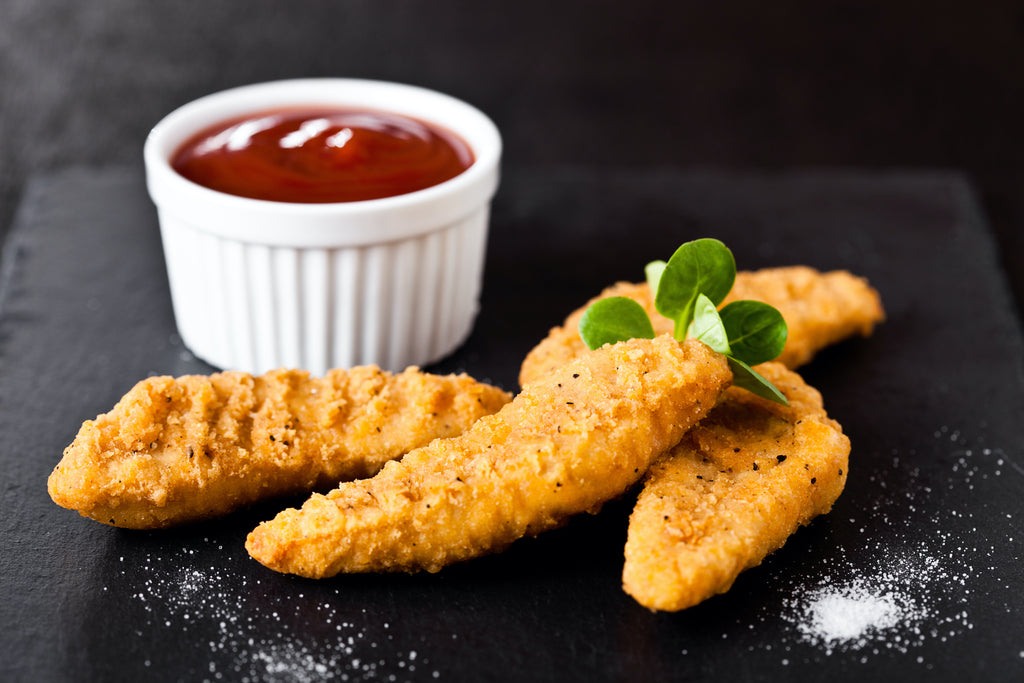 four cooked chicken tenders on a black slate board, with a small pot of ketchup
