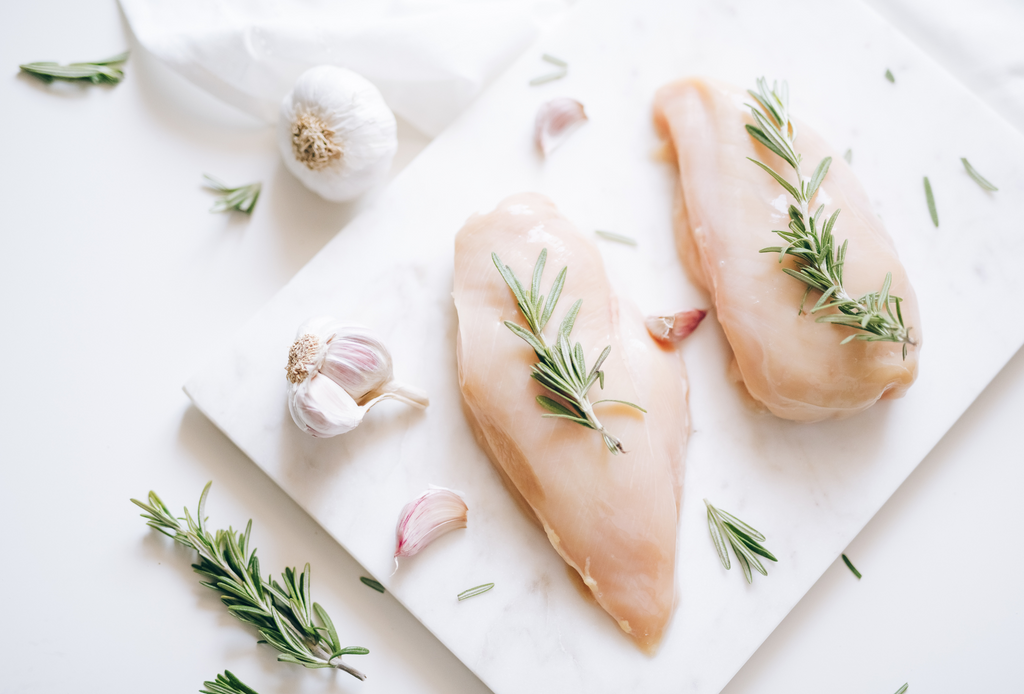 Two frozen chicken breasts surrounded by rosemary, and garlic bulbs on a marble platter