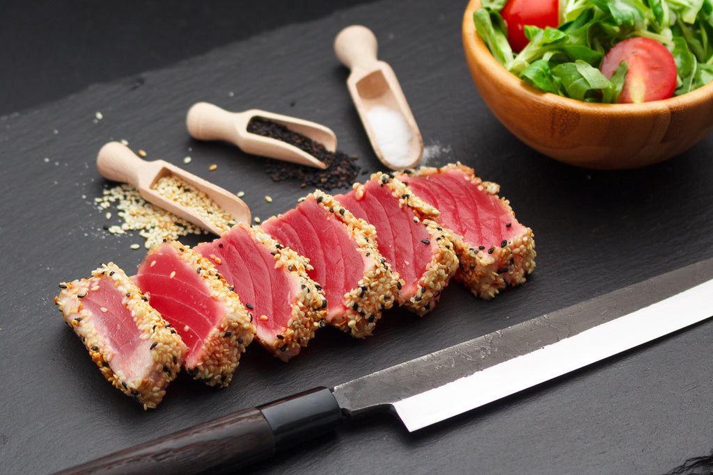 Six slices of tuna block surrounded by seasonings, butcher knife and a bowl of salad on a black cutting board