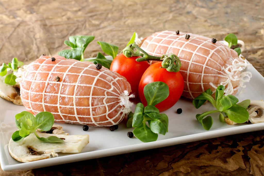 Two fresh, netted turkey rolls with thyme, tomatoes on a white plate against a beige background