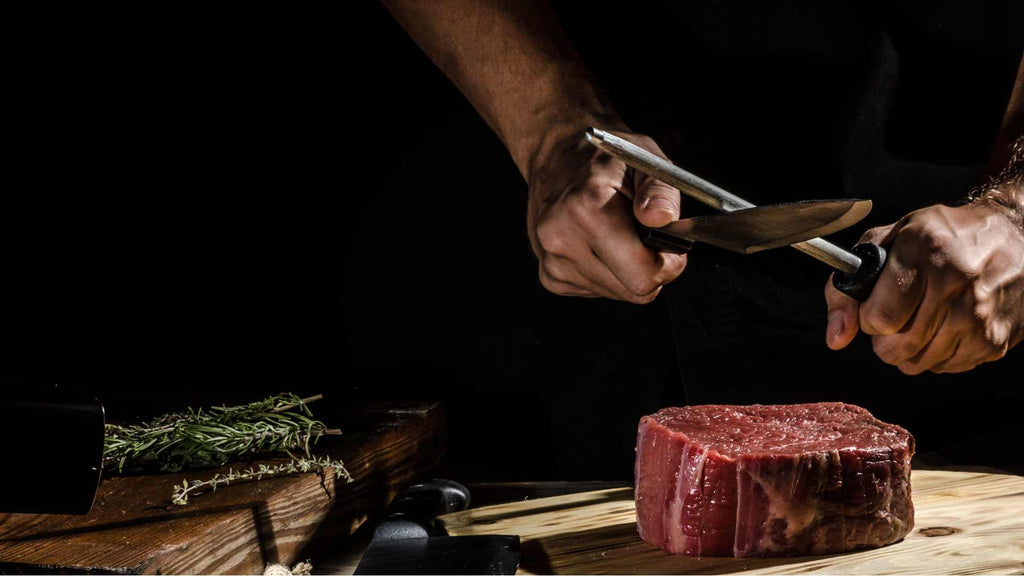 Butcher sharpening knives above a beef tenderloin steak on a wooden cutting board