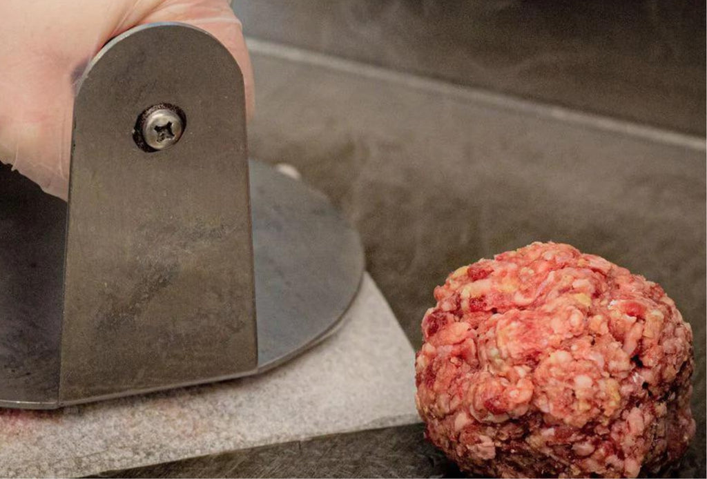 Fresh, seasoned meat ball beside a stainless steel smash burger press on a dark grey table