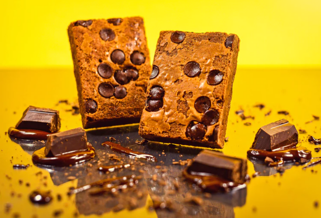 Two brownies embedded with chocolate chips surrounded with chunks of melted chocolate on a yellow background