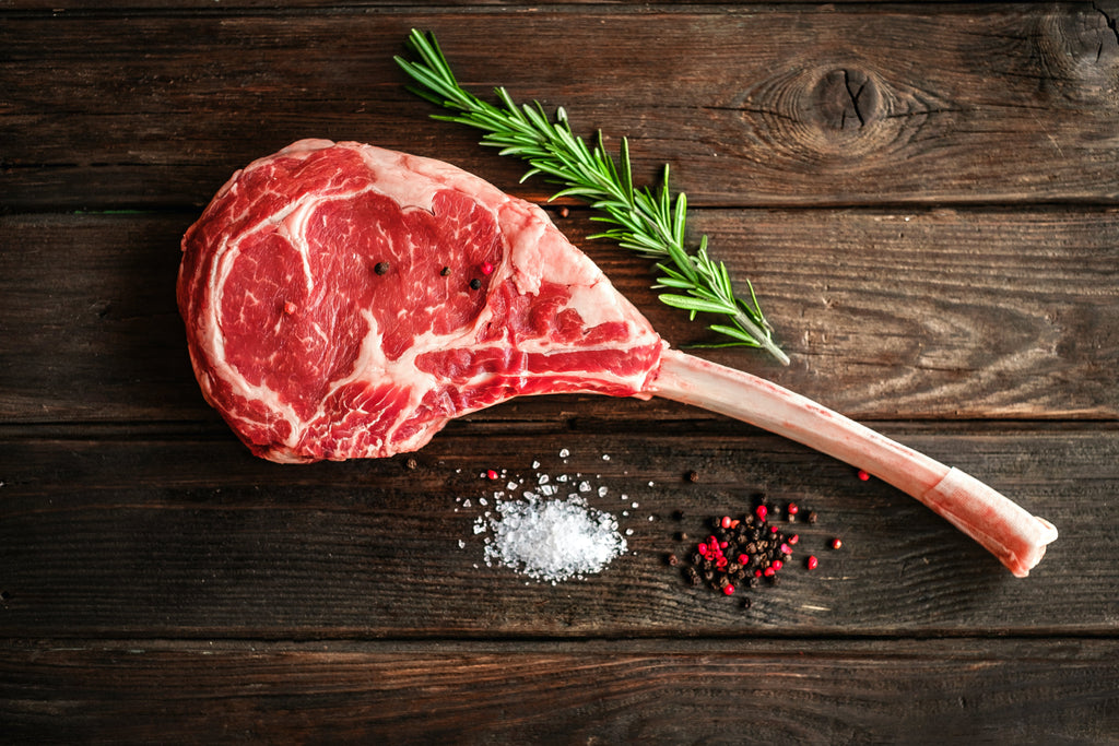 a tomahawk steak with a long bone sitting on a wooden board, with salt, peppercorns and rosemary sitting beside the steak