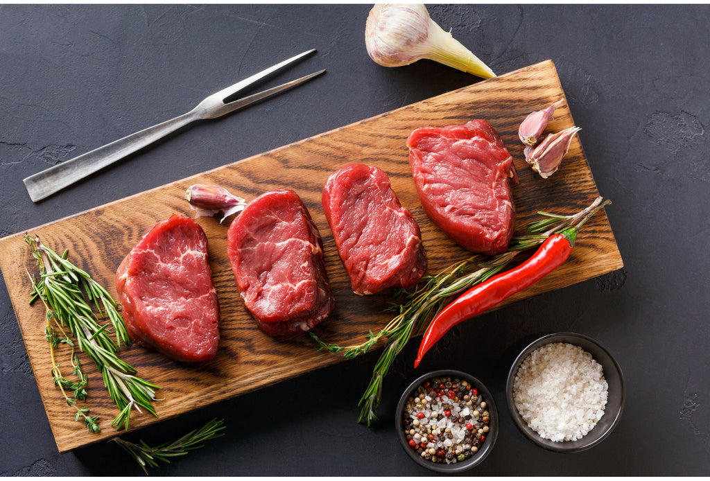 four uncooked beef tenderloin steaks on a wood platter with rosemary, garlic and seasoning, on a black background