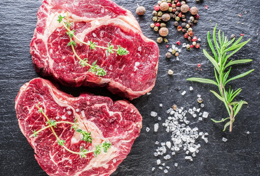 Two fresh ribeye steaks with peppercorns, sea salt and rosemary on a black slate table
