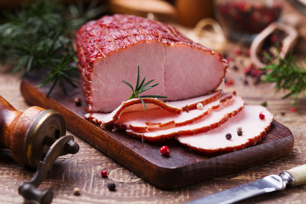 Sliced baked ham on wooden cutting block surrounded by peppercorns, rosemary and pepper grinder
