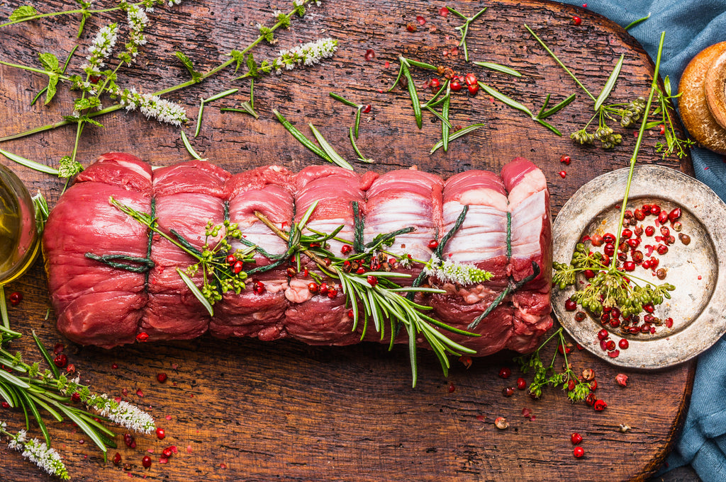 Fresh, uncooked AAA beef tenderloin surrounded by thyme and red peppercorns displayed on a wooden cutting board