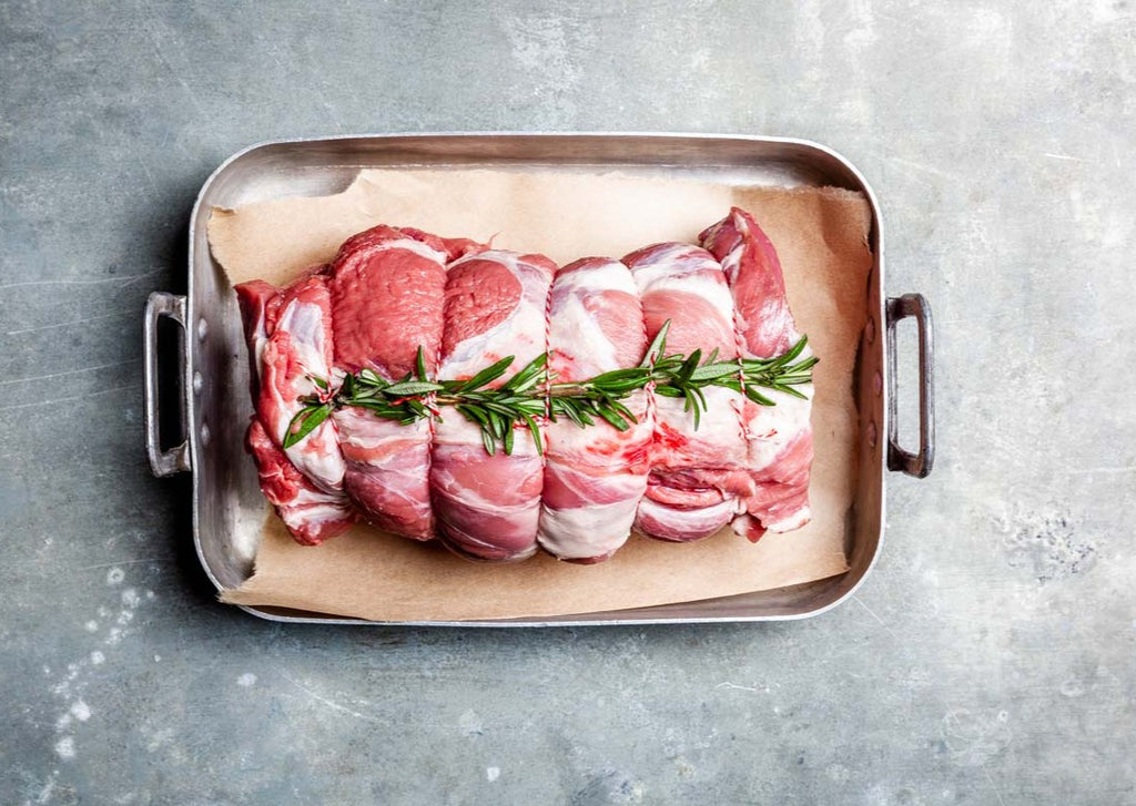 Fresh boneless lamb shoulder topped with rosemary lying on brown parchment paper in a silver roasting pan