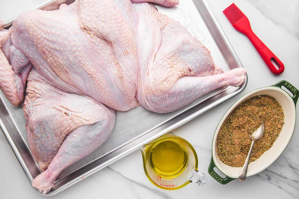 Fresh flattened turkey on a silver roasting pan surrounded by olive oil, fresh seasonings and a red basting brush