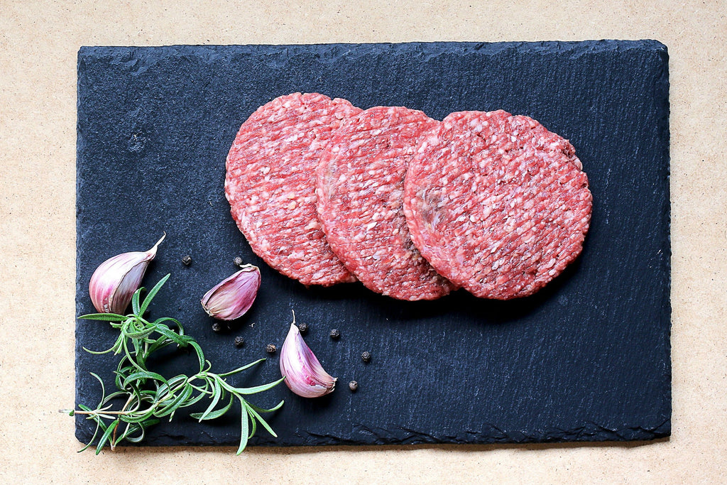 Three fresh ground beef burger patties beside garlic bulbs and thyme displayed on a black slate cutting board