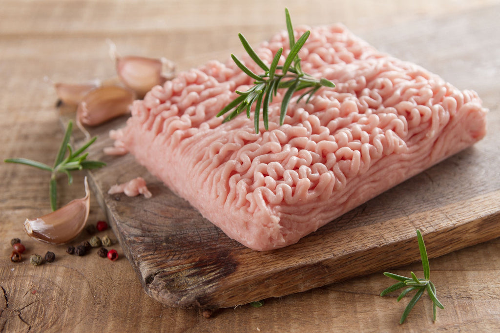 A selection of fresh ground chicken surrounded by garlic bulbs and peppercorns on a distressed wooden cutting board
