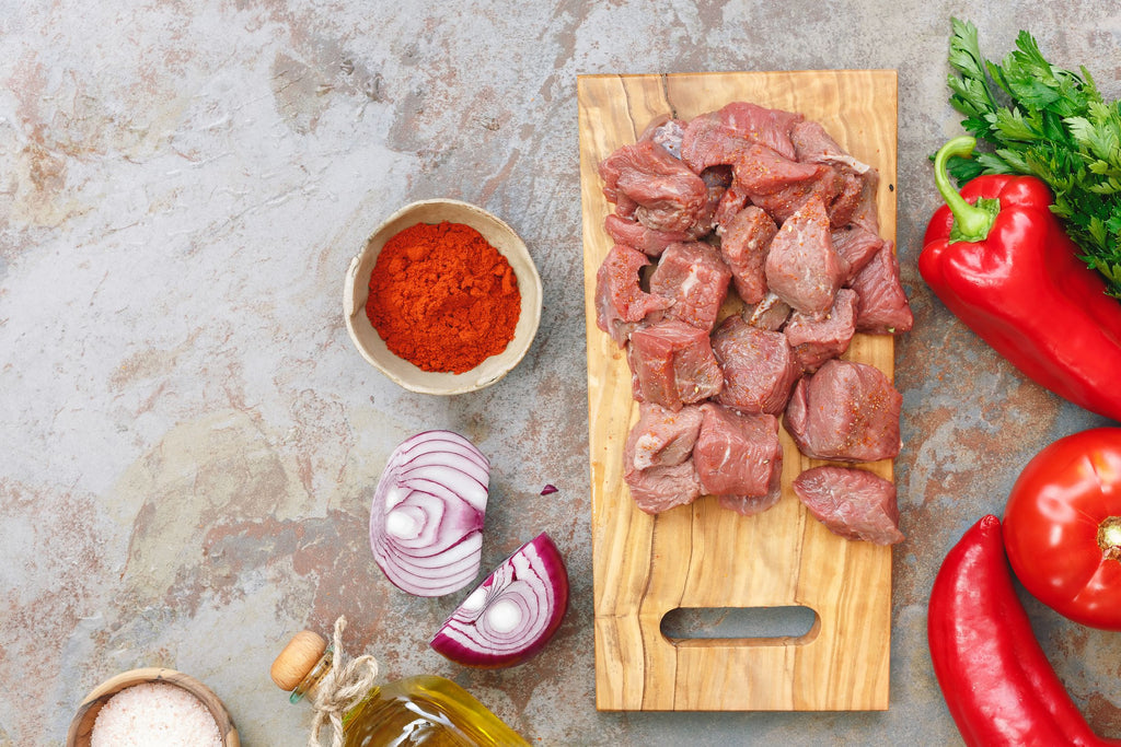 Lamb stew cubes on a cutting board surrounded by red peppers, red onions against a marble background