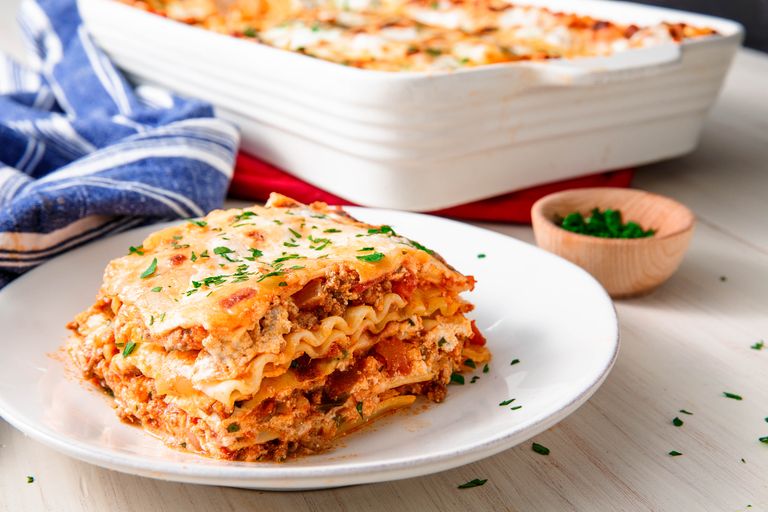Cooked portion of meat lasagna on a white serving saucer dish beside a large lasagne cooking dish