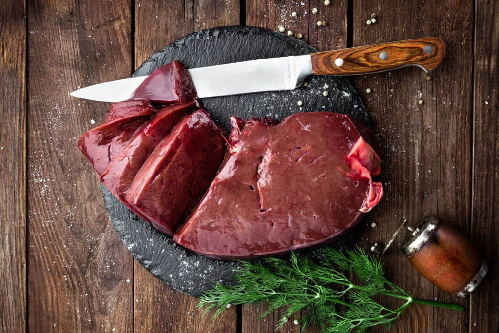 A selection of fresh liver sliced beside a butcher knife on a black slate cutting board beside rosemary and pepper mill.