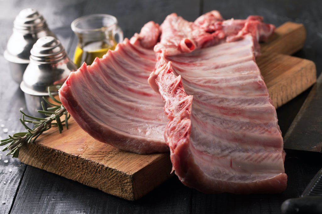 two pork baby back ribs on a wooden cutting board, with seasonings and herbs in background
