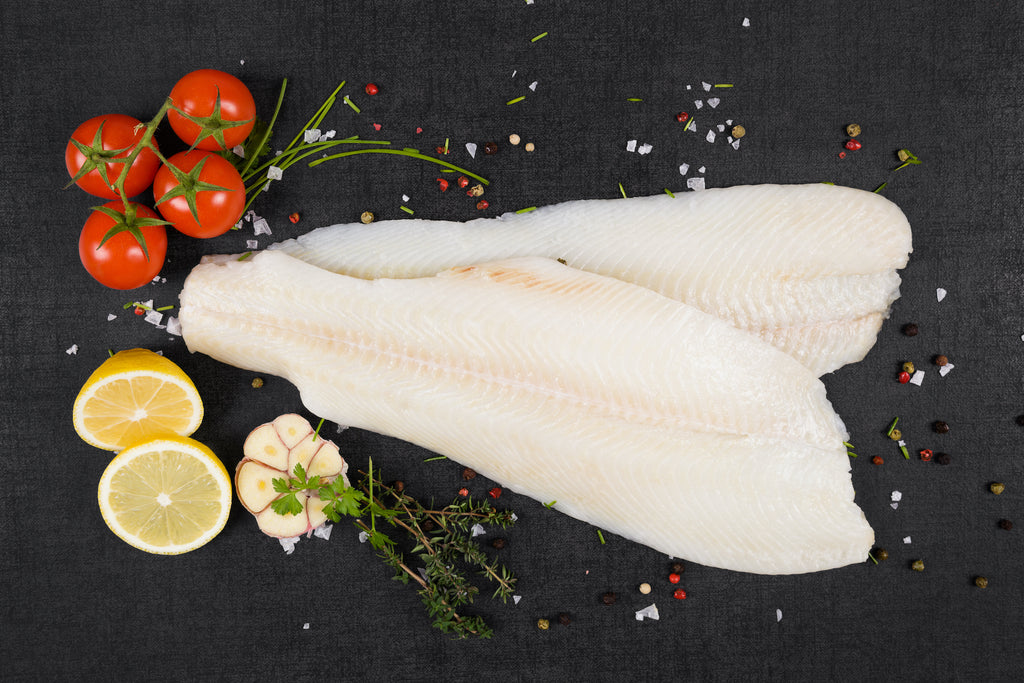 two filets of fresh halibut, surrounded by cherry tomatoes, lemon, garlic and seasonings, on a black slate background