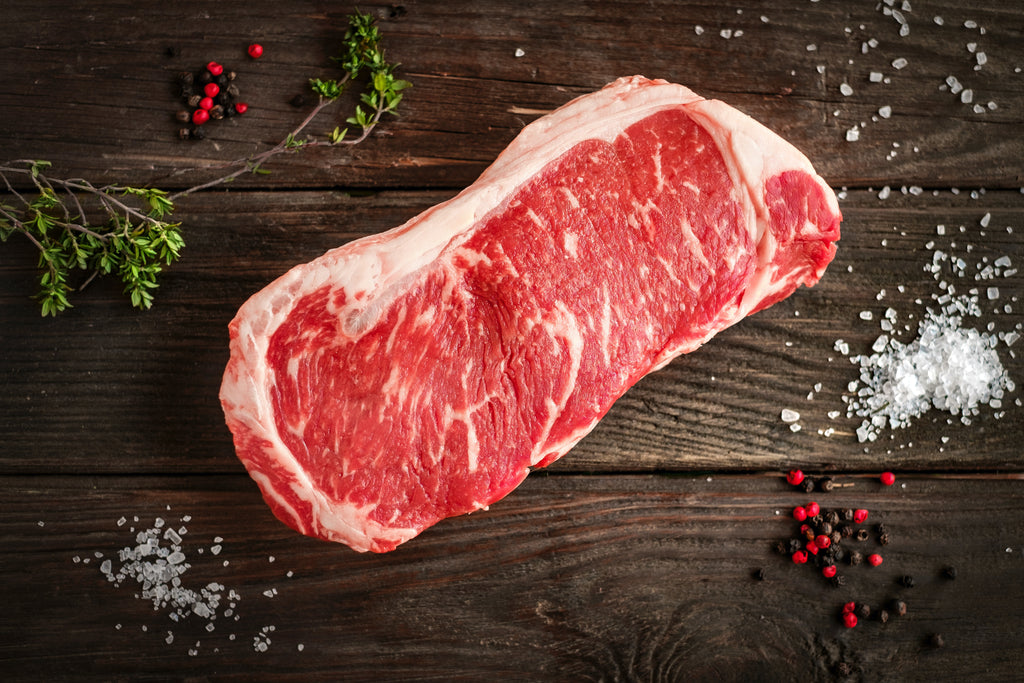 A fresh center-cut of striploin steak surrounded by peppercorns, sea salt and thyme on a black wooden background